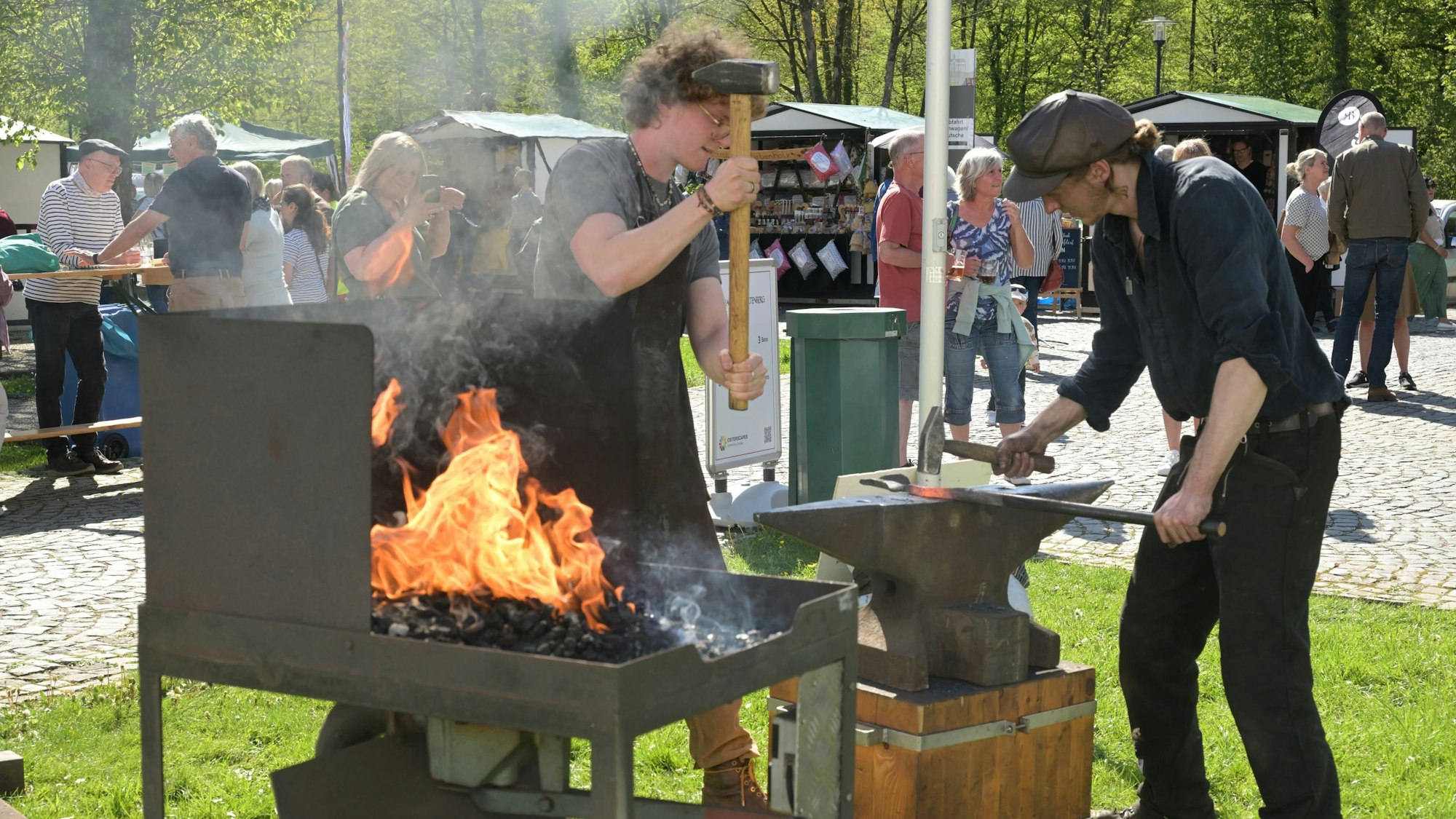 Zwei Personen hauen mit Hämmern auf eine Eisenstange, die auf einem Amboss liegt.