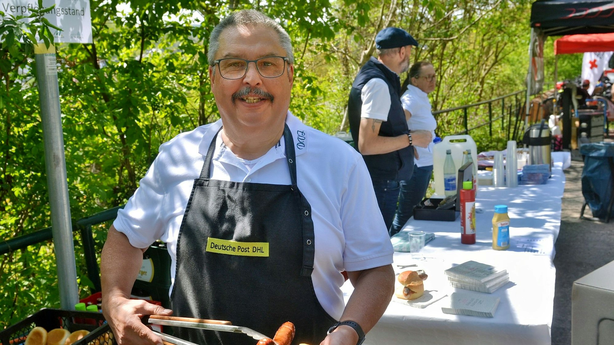 Das Bild zeigt Wilfried Schumacher mit Grillzange und Bratwurst vor einem Verkaufsstand.