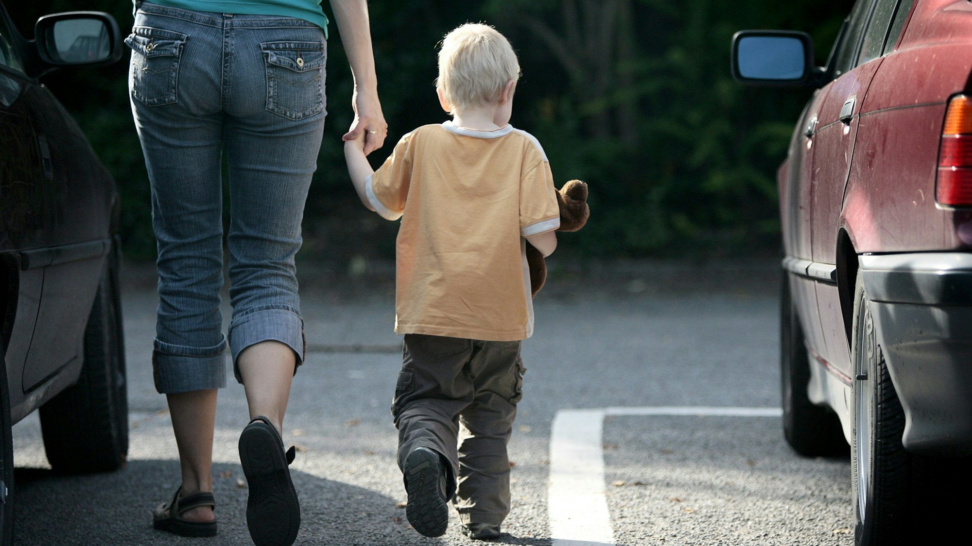 Eine Mutter geht mit ihrem Sohn an der Hand zwischen Autos durch. Das Kind hat ein Kuscheltier im Arm.