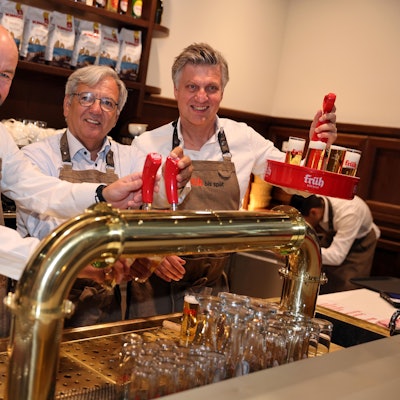 Drei Männer in beiger früh Kölsch Schürze, lächeln und stehen hinter einer Theke, zapfen Kölsch, einer der Männer hält einen Kranz hoch.