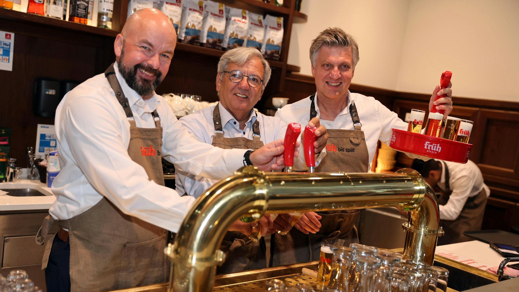 Drei Männer in beiger früh Kölsch Schürze, lächeln und stehen hinter einer Theke, zapfen Kölsch, einer der Männer hält einen Kranz hoch.