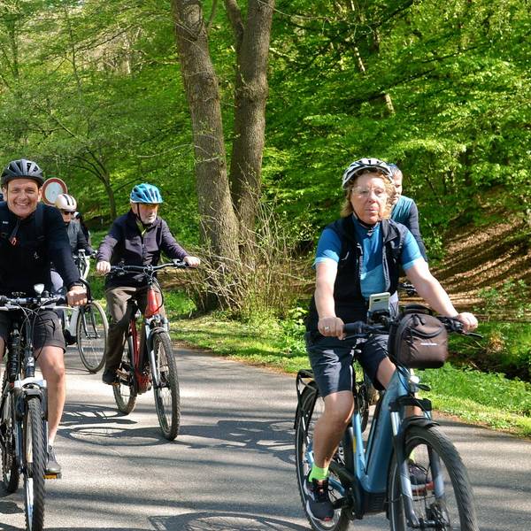Das Bild zeigt Radfahrerinnen und Radfahrer auf einem asphaltierten Weg, im Hintergrund Bäume und Sträucher.