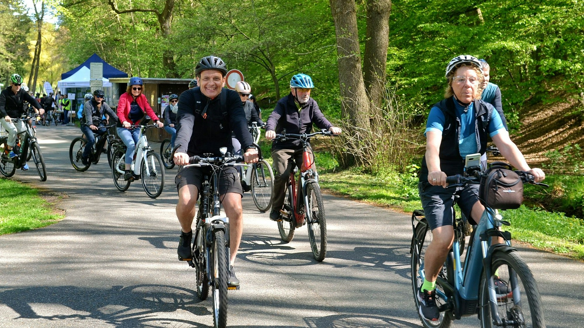 Das Bild zeigt Radfahrerinnen und Radfahrer auf einem asphaltierten Weg, im Hintergrund Bäume und Sträucher.
