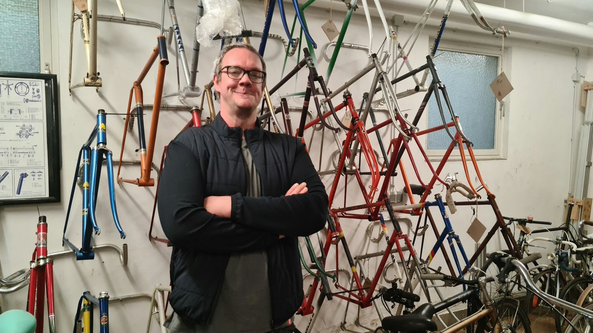Ein Mann mit Brille und dunklen Klamotten steht vor einer Wand, an der Fahrradrahmen in allen Farben hängen.