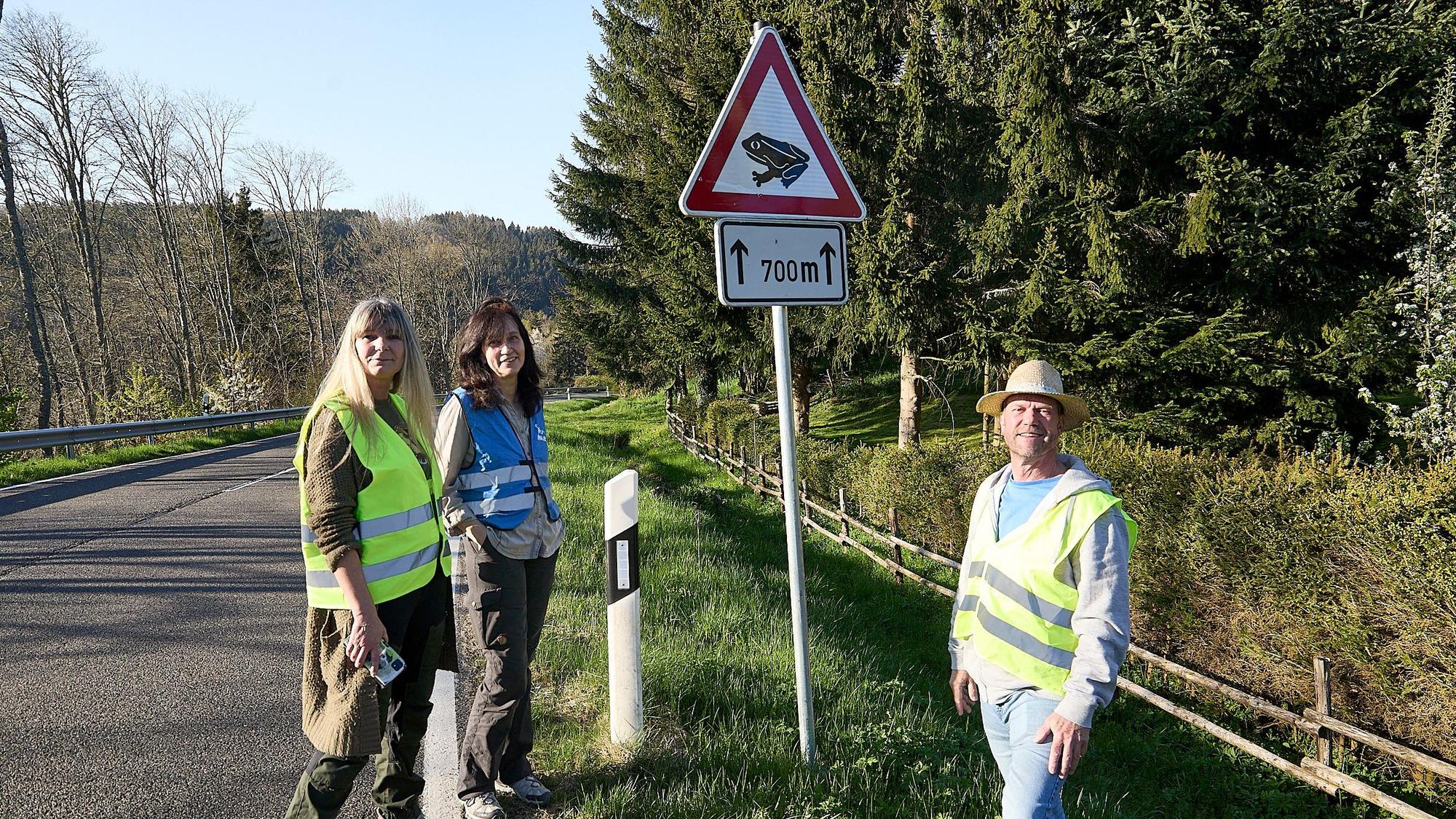 Zwei Frauen und ein Mann tragen Warnwesten. Sie stehen an der Landstraße bei Hellenthal-Wildenburg, an der ein Warnschild mit einer Kröte angebracht ist, das auf die Wanderung der Amphibien hinweist.