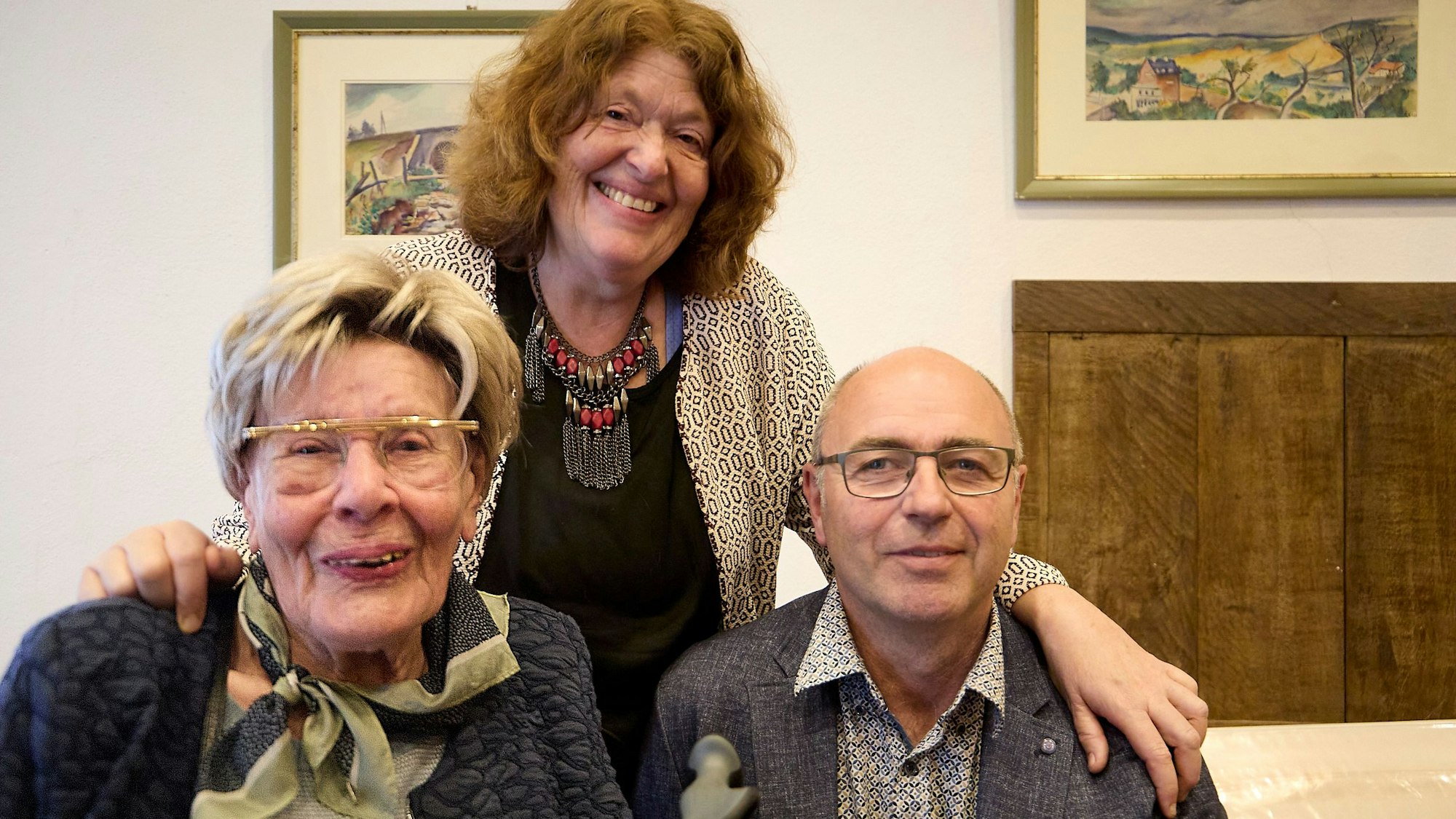 Ein Mann und eine Frau sind im Vordergrund zu sehen. Hinter ihnen steht eine Frau, die beiden die Hände auf die Schulter legt.