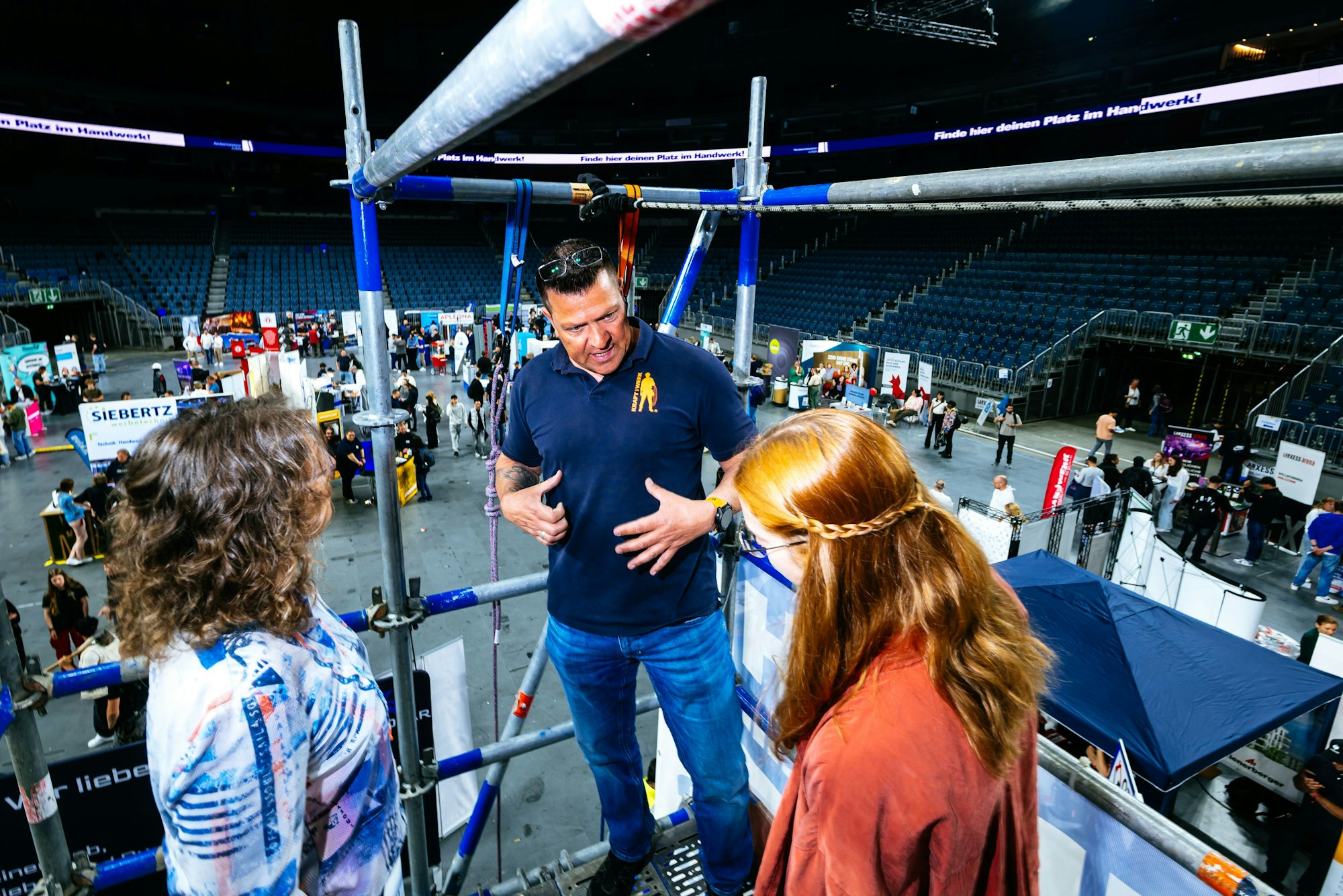 Ein Mann steht mit zwei Frauen auf einem Baugerüst in der Arena und erklärt etwas. Hinter ihnen sieht man weitere Messestände und Menschen.