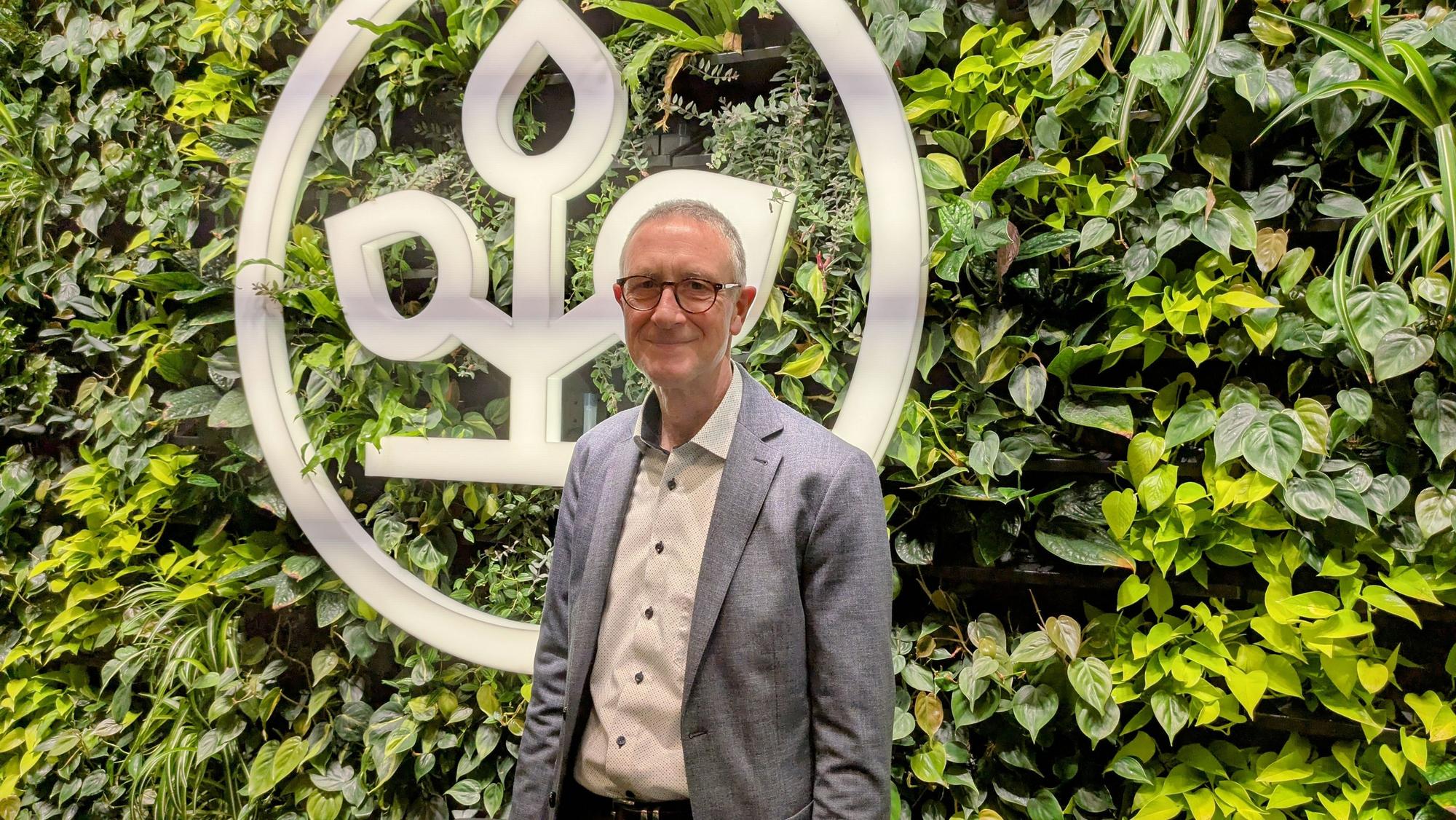 Helmut Schneider steht vor einer begrünten Wand mit einem AOK-Emblem.