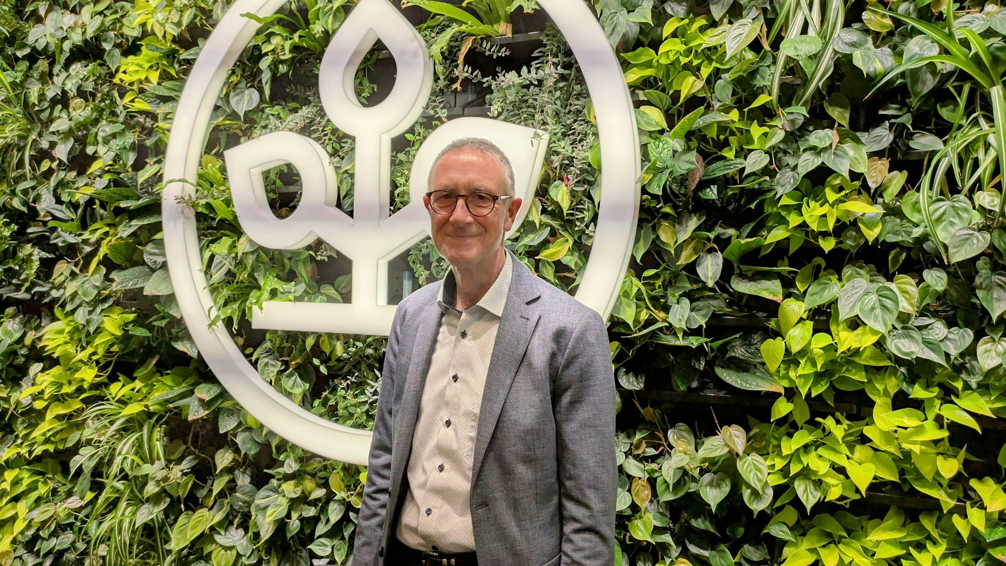 Helmut Schneider steht vor einer begrünten Wand mit einem AOK-Emblem.