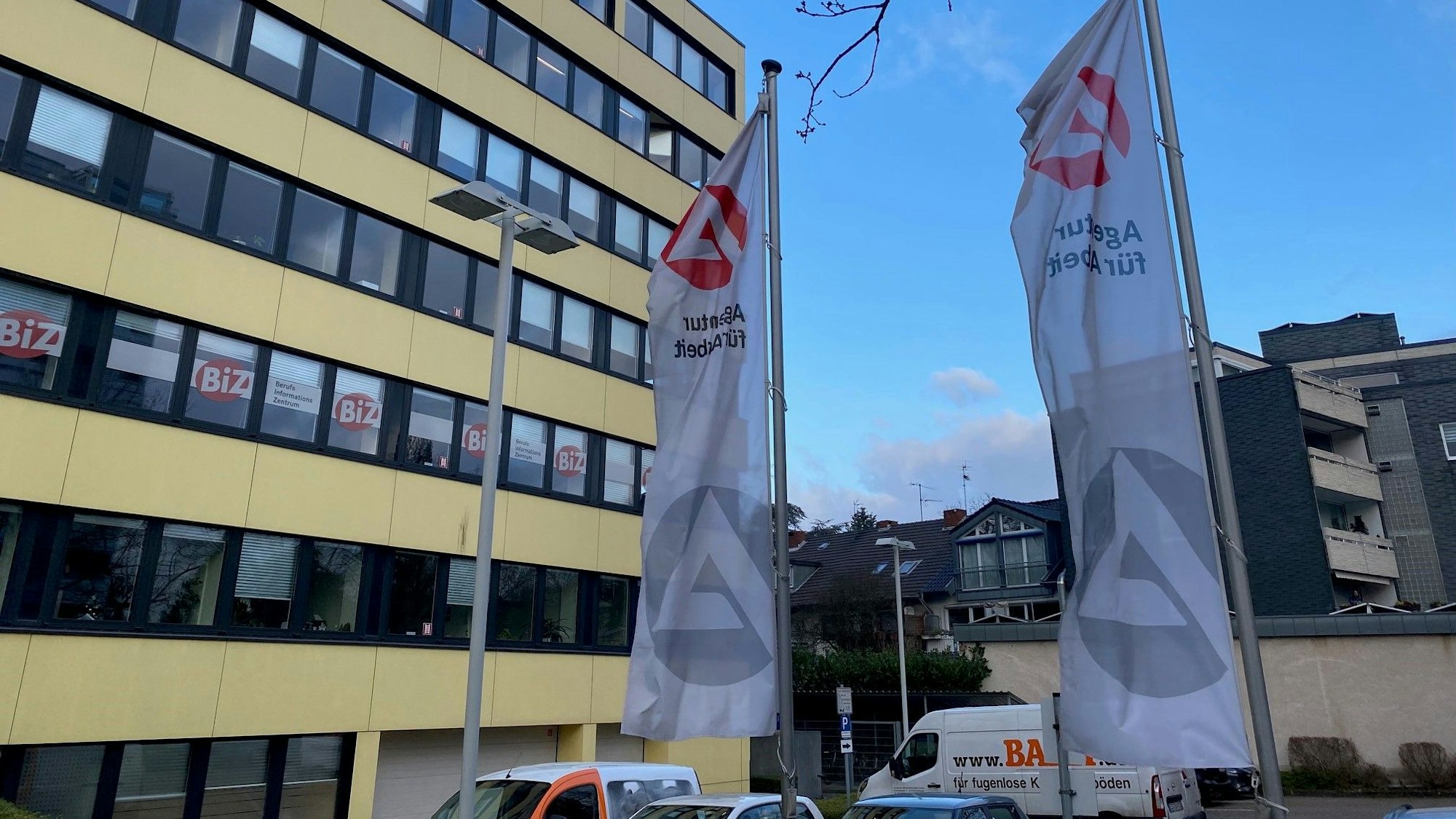 Zu sehen ist das Gebäude der Agentur für Arbeit in Brühl, vor dem zwei Fahnen mit dem Logo der Behörde wehen.