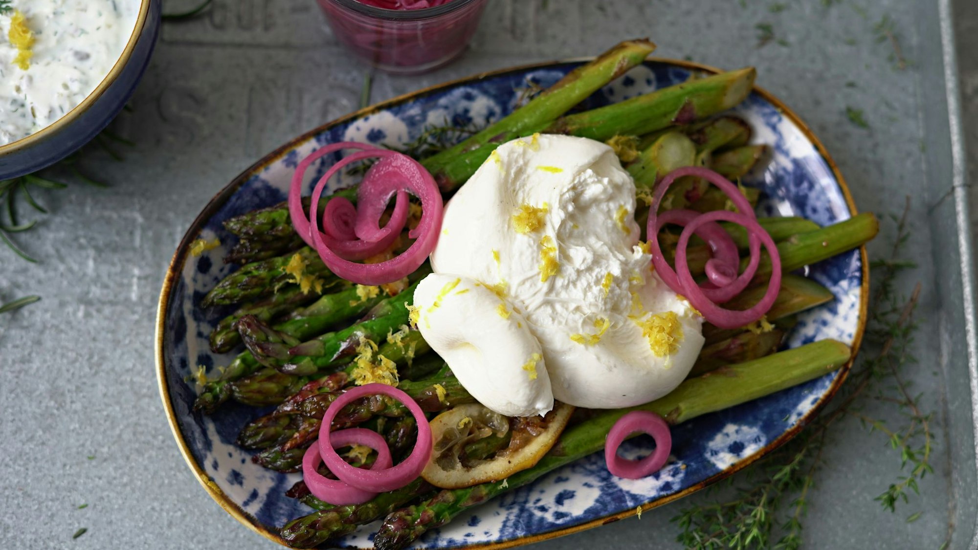 Zum Anrichten wird der Burrata über den Spargelstangen etwas auseinander gezupft und mit dem Ranchdip beträufelt. Zuvor eingelegte rote Zwiebelringe bringen Farbe auf den Teller.
