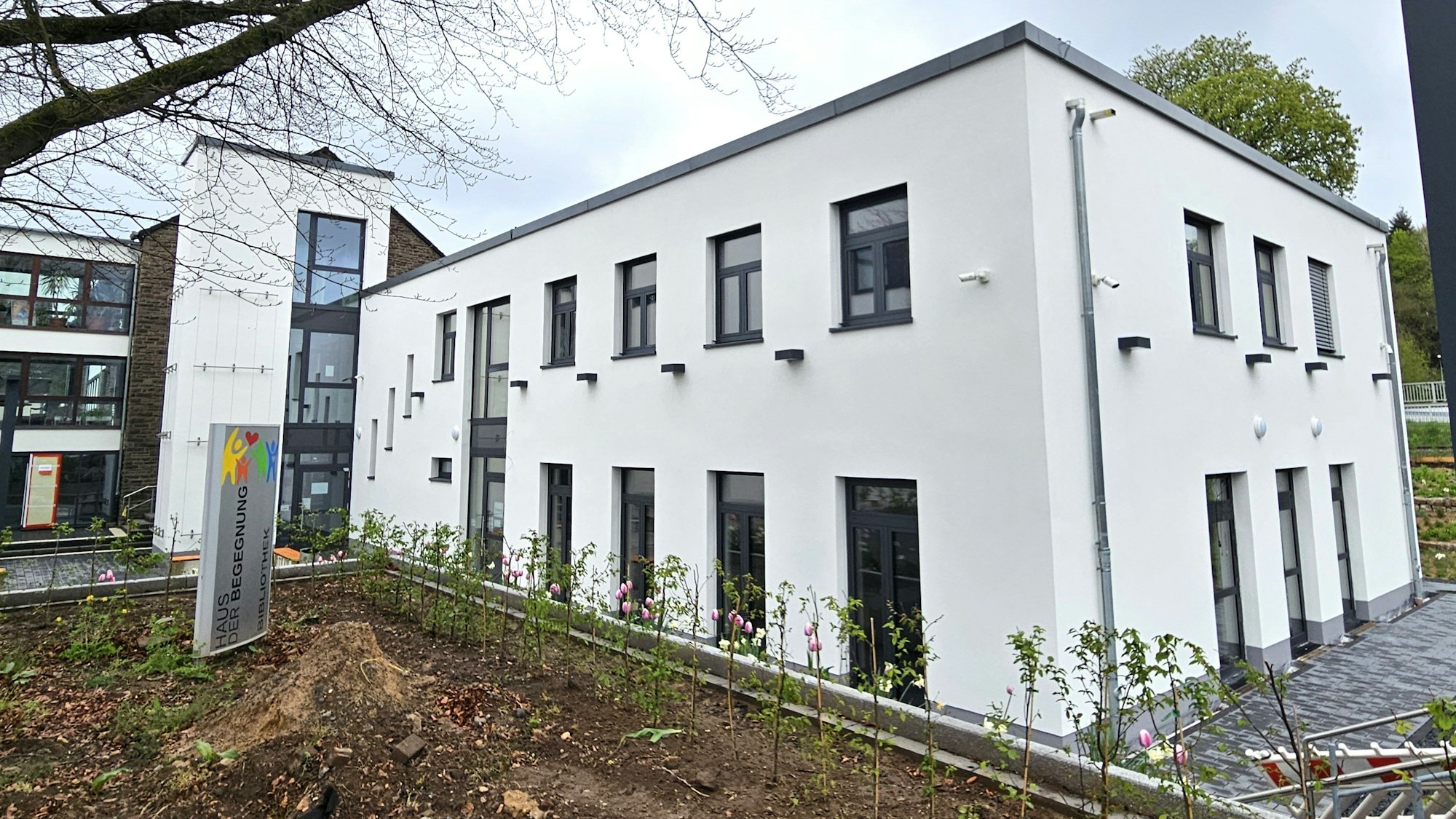 Ein weißer rechteckiger Neubau steht neben einem Beet und einer frisch gepflanzten Hecke.