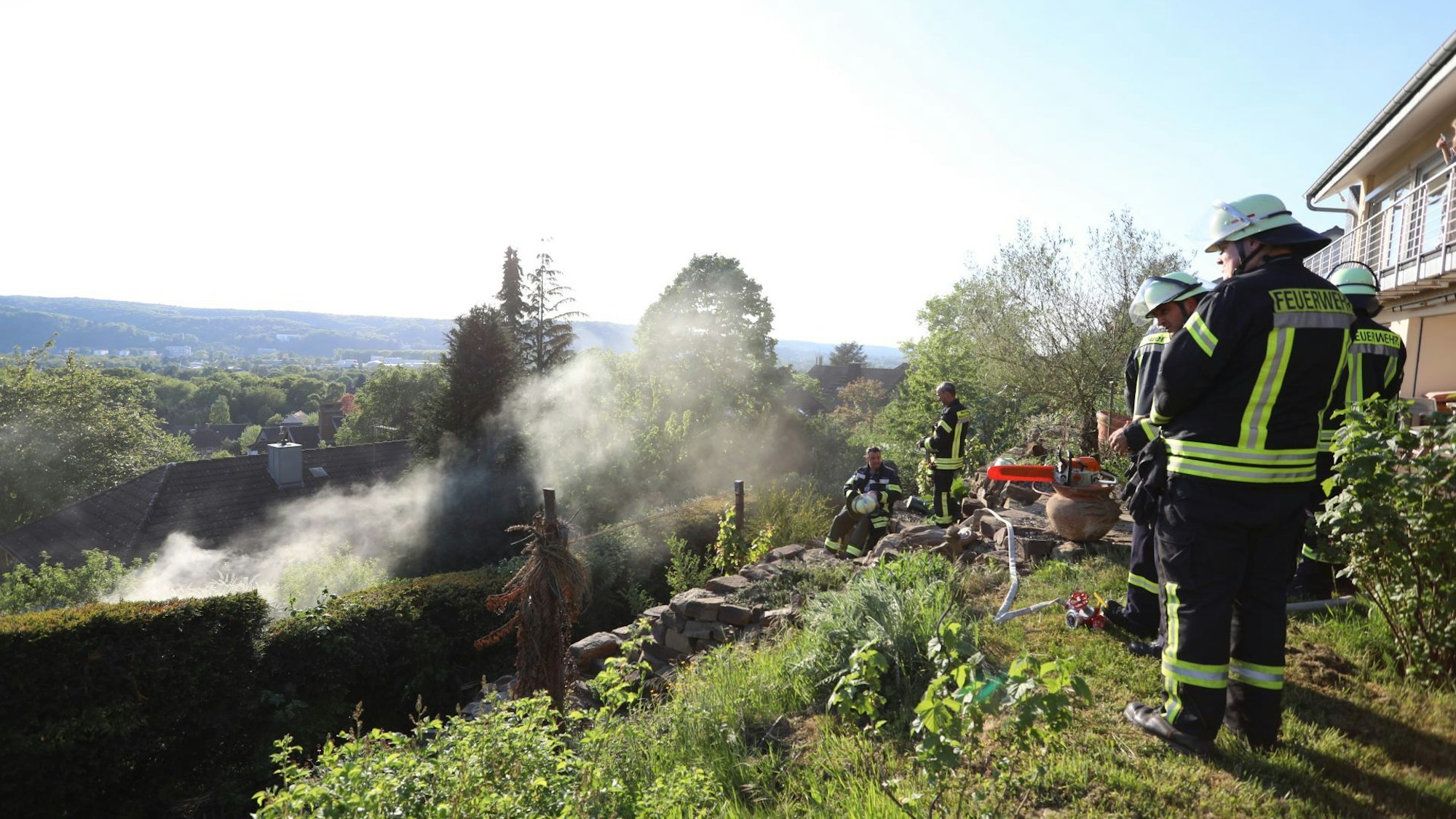 Feuerwehrleute stehen auf einem Grundstück oberhalb der Stadt, ein Stück weiter unten steigt Rauch auf.