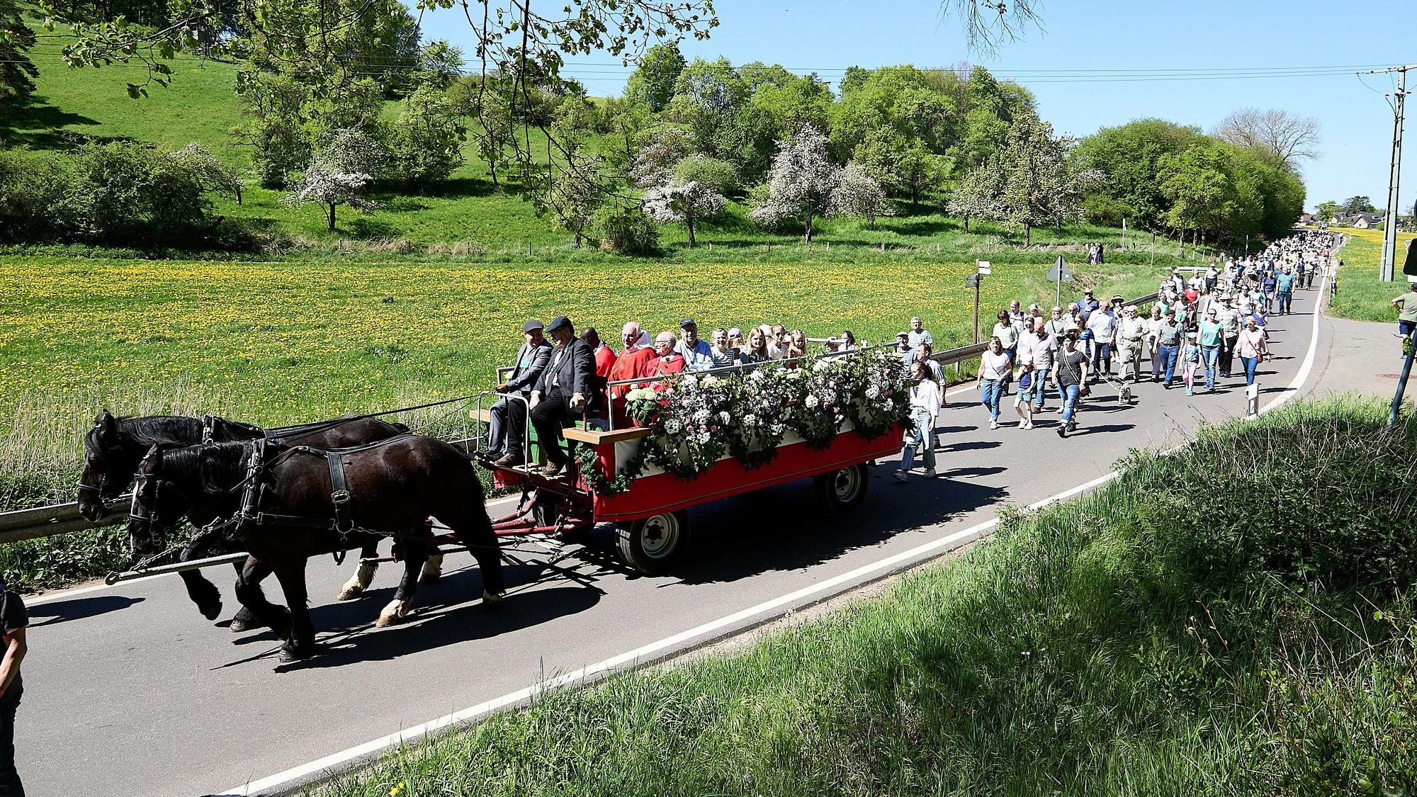 Ein roter Wagen, der von zwei schwarzen Pferden gezogen wird, zieht vor einem Zug Menschen über eine Straße, drumherum sind grüne Hügel und Bäume zu sehen.