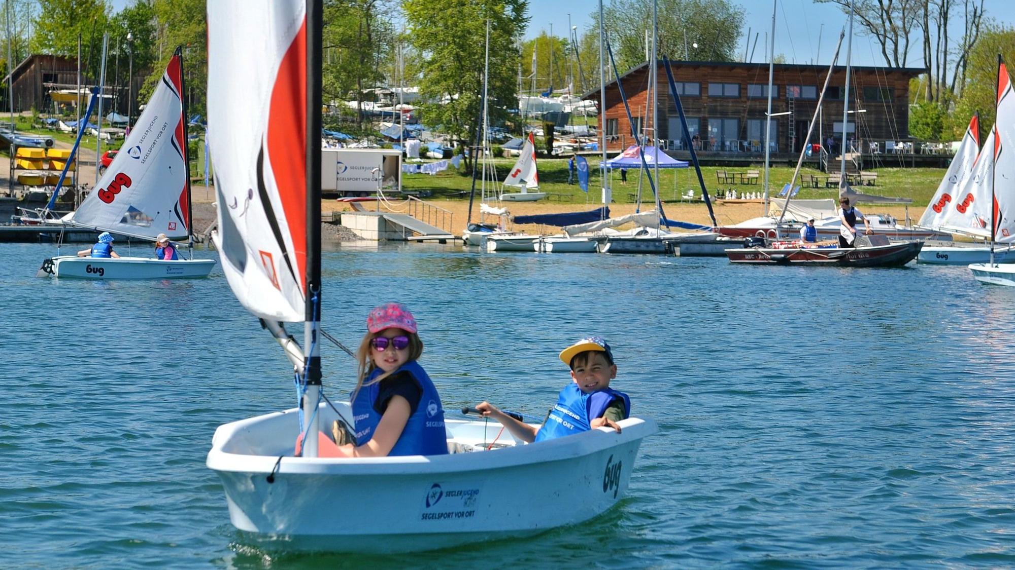Zwei Kinder segeln über den Wassersportsee, im Hintergrund ist das Ufer zu sehen.