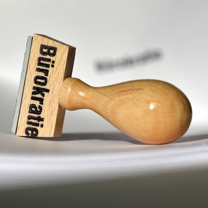 Ein Stempel mit der Aufschrift „Bürokratie“ liegt auf Papieren.