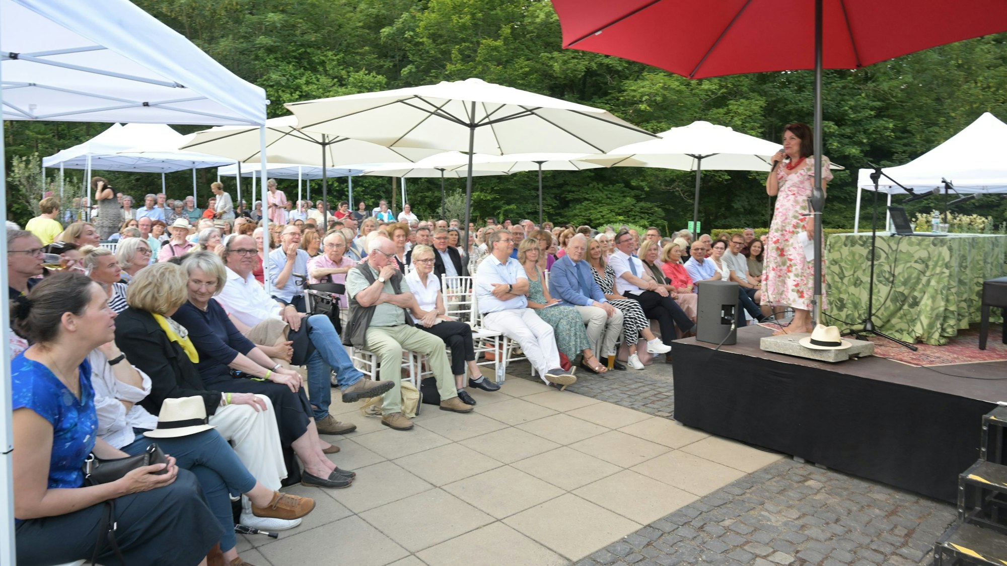Viele Menschen sitzen unter Sonnenschirmen vor einer Bühne. Darauf einer Frau im Sommerkleid am Mikrofon.
