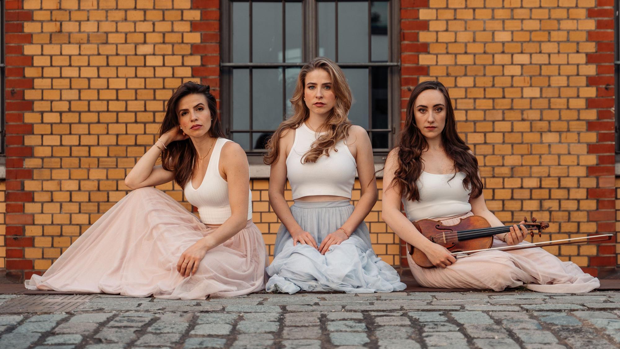 Drei Frauen, auf dem Boden sitzend. Die, die rechts sitzt, hält eine Geige und einen Geigenbogen in der Hand.