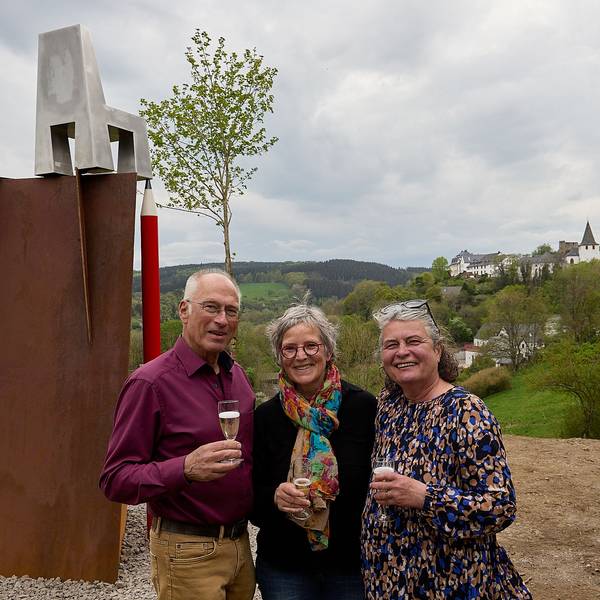 Ein Mann und zwei Frauen halten jeder ein Glas Sekt in der Hand. Links von ihnen ist eine neue Skulptur zu sehen, rechts der Blick auf den Ort Kronenburg.