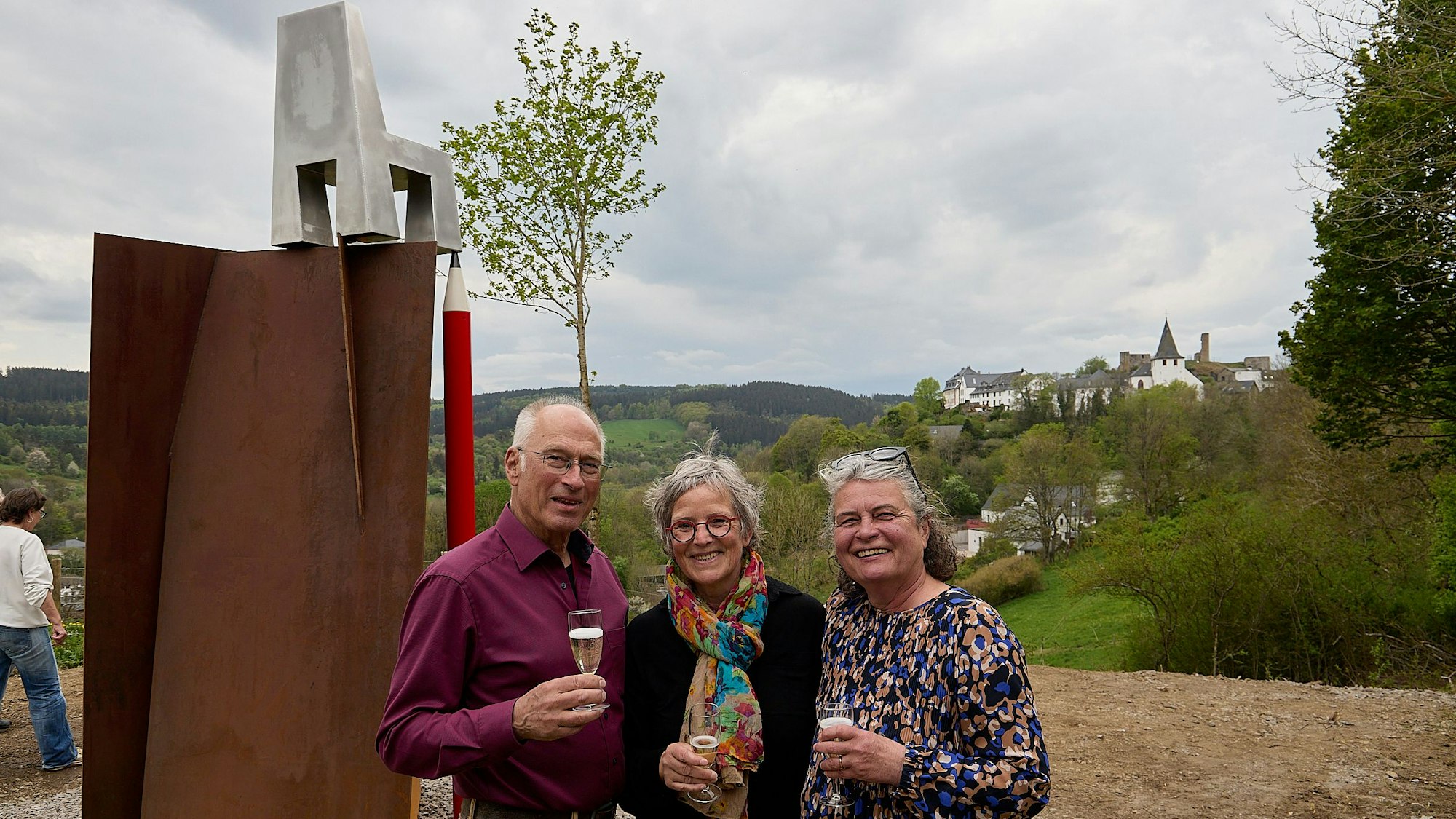 Ein Mann und zwei Frauen halten jeder ein Glas Sekt in der Hand. Links von ihnen ist eine neue Skulptur zu sehen, rechts der Blick auf den Ort Kronenburg.