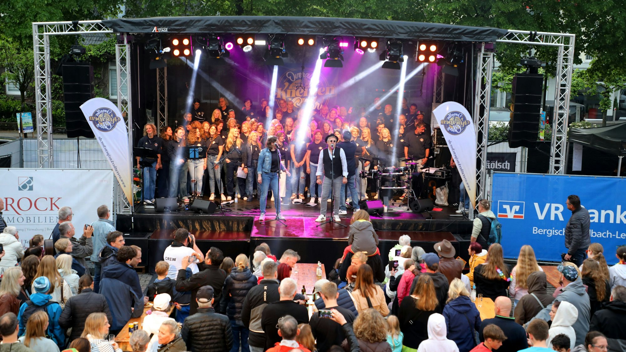 Zu sehen ist eine bunt beleuchtete Bühne, mit dem Auftritt des Overather Kneipenchors. Davor sammelt sich eine Menge Publikum.