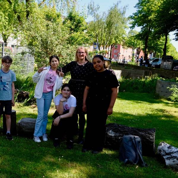 Zwei Jungs, drei Mädchen und eine Frau mit langem blondem Haar stehen in einem Garten.
