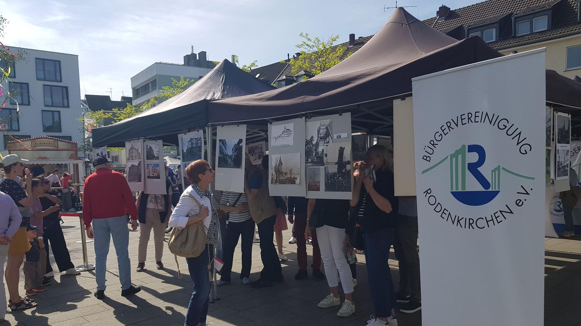 Menschen stehen vor Bildern, die unter einem Pavillon auf einem Platz aufgehängt wurden.