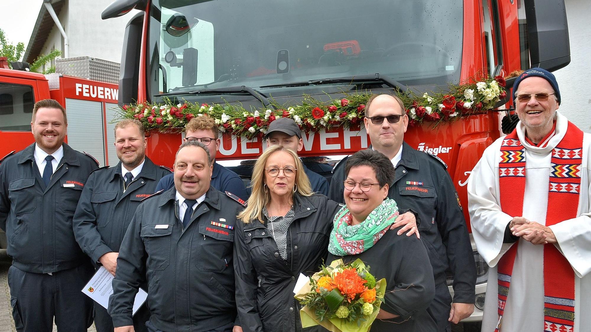 Eine Gruppe Männer und Frauen steht vor einem mit einer Blumengirlande geschmückten neuen Feuerwehrfahrzeug.
