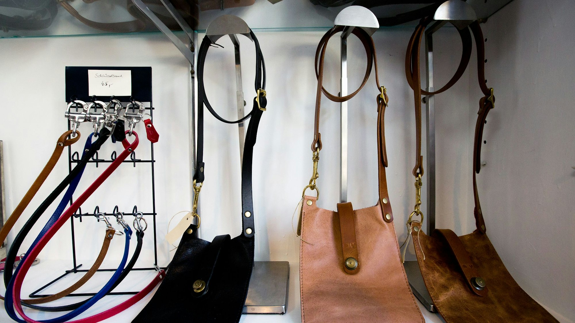 Hundeleinen und Schlüsselanhänger hängen an einem Gestell, daneben sind drei Ledertaschen in Schwarz und Brauntönen ausgestellt.