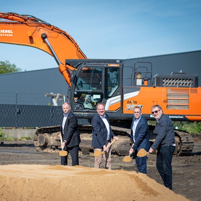 Das Foto zeigt vier Männer mit Spaten vor einem Sandhaufen, im Hintergrund ein Bagger.