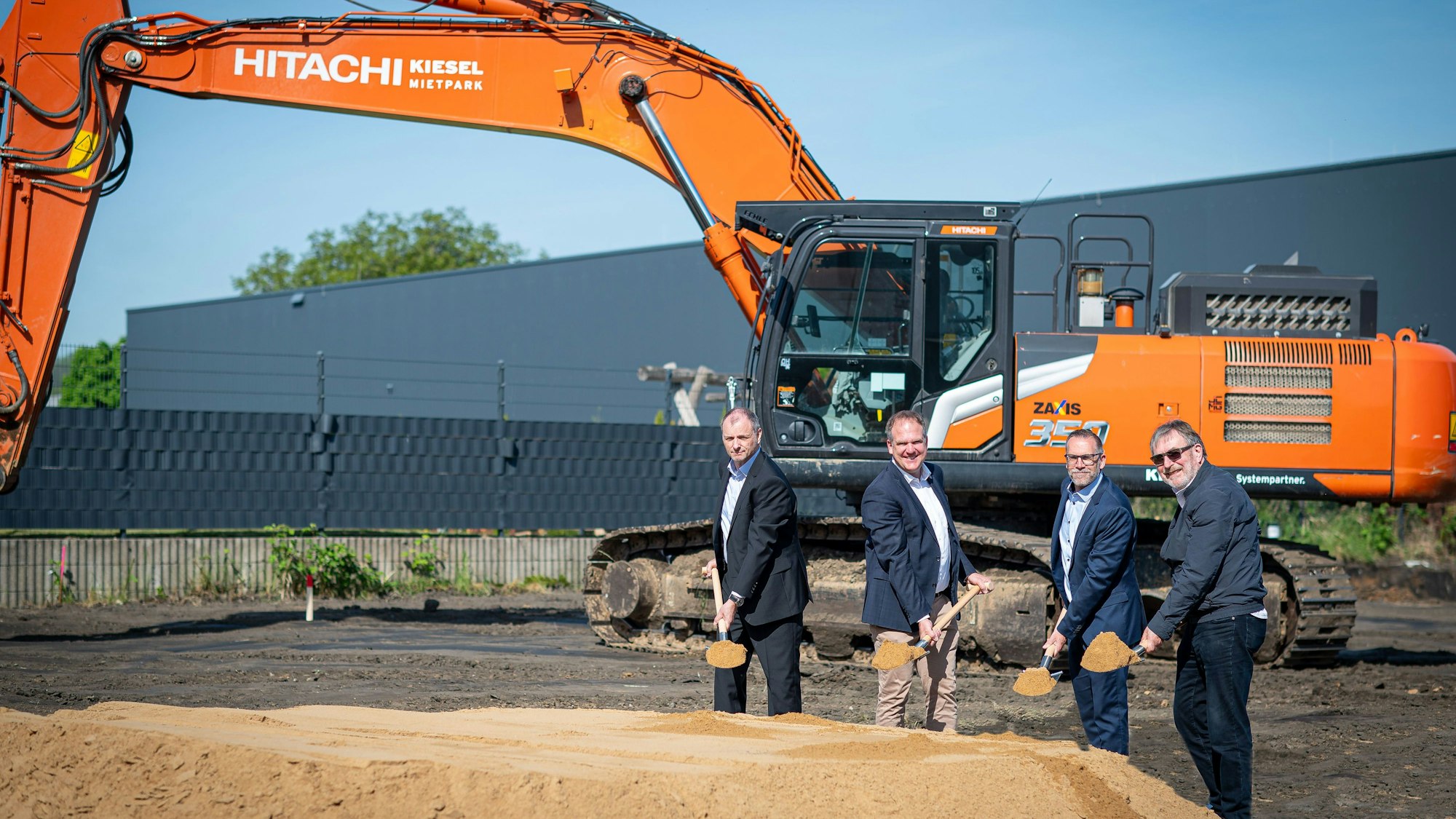 Das Foto zeigt vier Männer mit Spaten vor einem Sandhaufen, im Hintergrund ein Bagger.