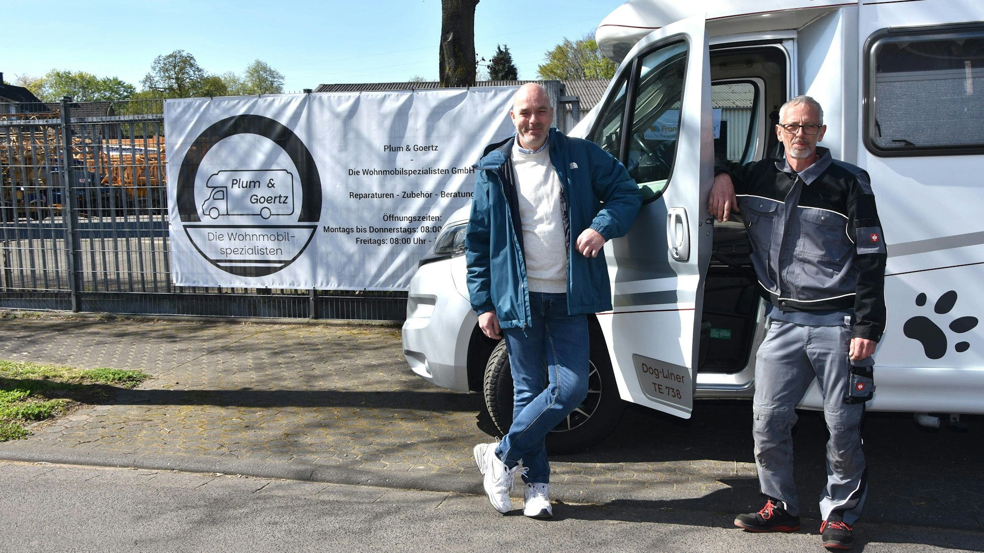 Auf dem Bild stehen die beiden Männer, angelehnt an die Tür eines Wohnmobils. Im HIntergrund an einem Zaun ein Banner mit dem Firmenlogo, einem Wohnmobil mit der Aufschrift Plum & Goertz.