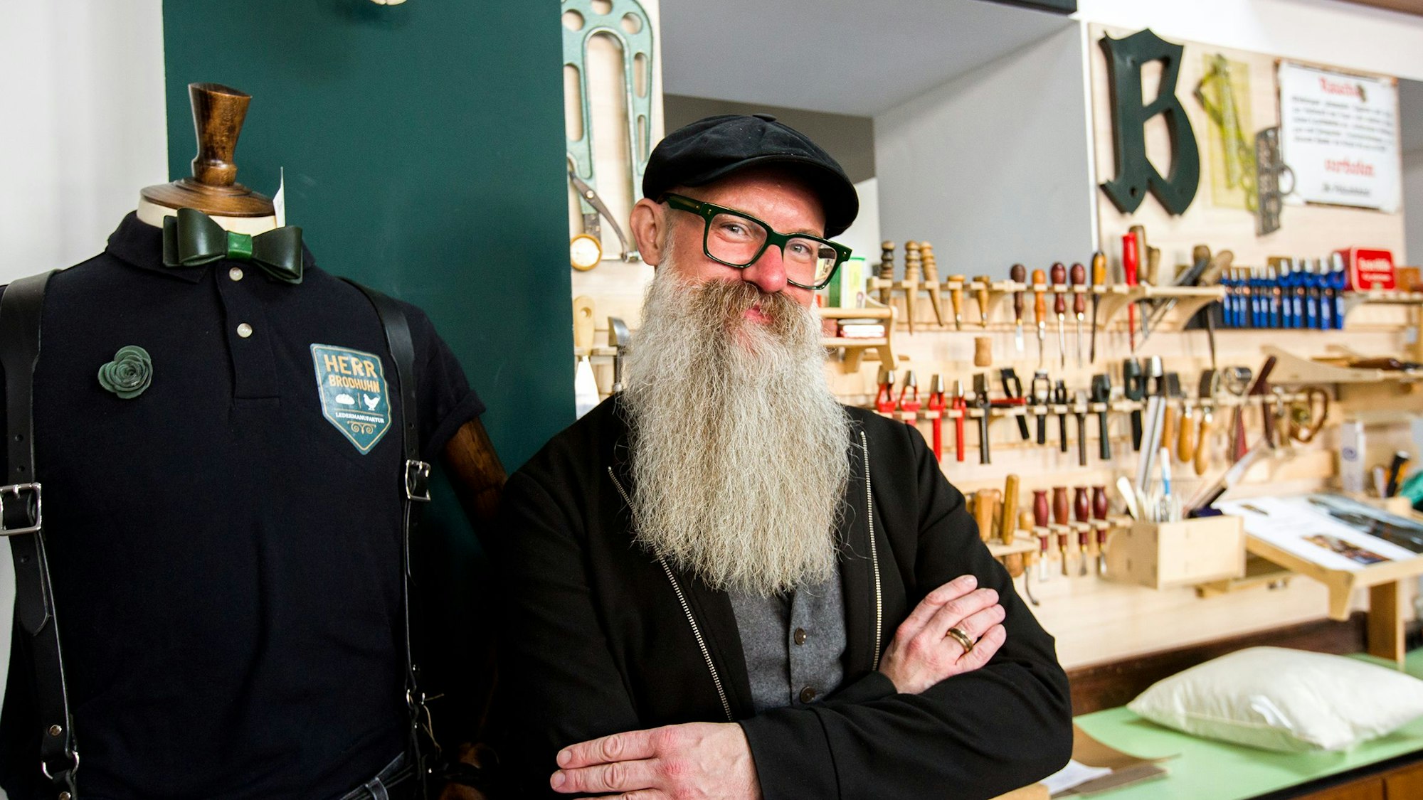 Ein Mann mit Brille und langem grauen Bart steht mit verschränkten Armen vor einer Werkbank mit allerlei Werkzeugen.