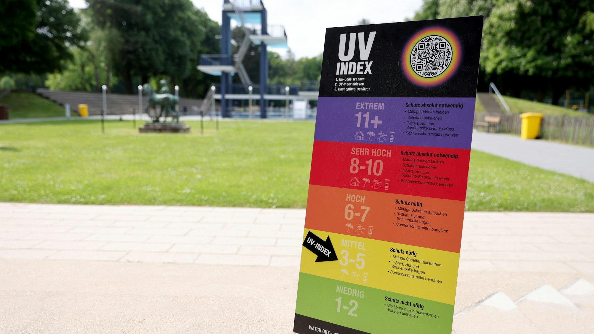 Im Hintergrund sieht man eine Grünfläche und den 10-Meter Turm des Freibads. Vorne steht die UV-Index-Tafel mit bunten Streifen, wobei auf jedem Streifen die jeweiligen Zahlen und Informationen geschrieben sind.