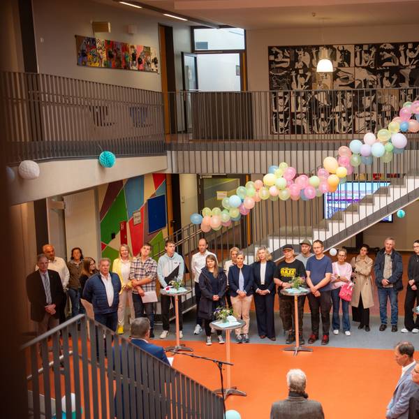Einige Menschen stehen in einem mit Blumen und Luftballons geschmückten Foyer und hören einem Redner zu.