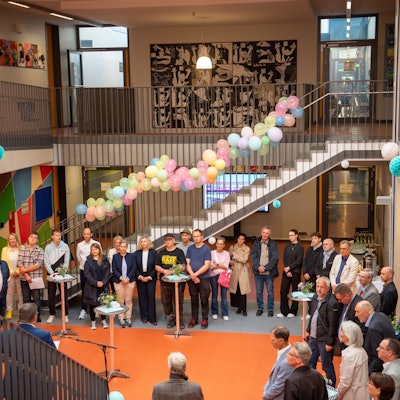 Einige Menschen stehen in einem mit Blumen und Luftballons geschmückten Foyer und hören einem Redner zu.