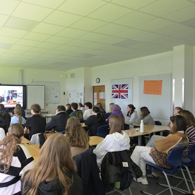 Schüler sitzen in ihrem Klassenzimmer und folgen einer Videokonferenz auf der großen Leinwand.