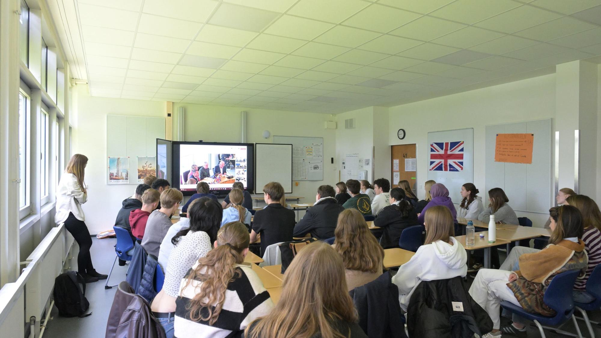 Schüler sitzen in ihrem Klassenzimmer und folgen einer Videokonferenz auf der großen Leinwand.