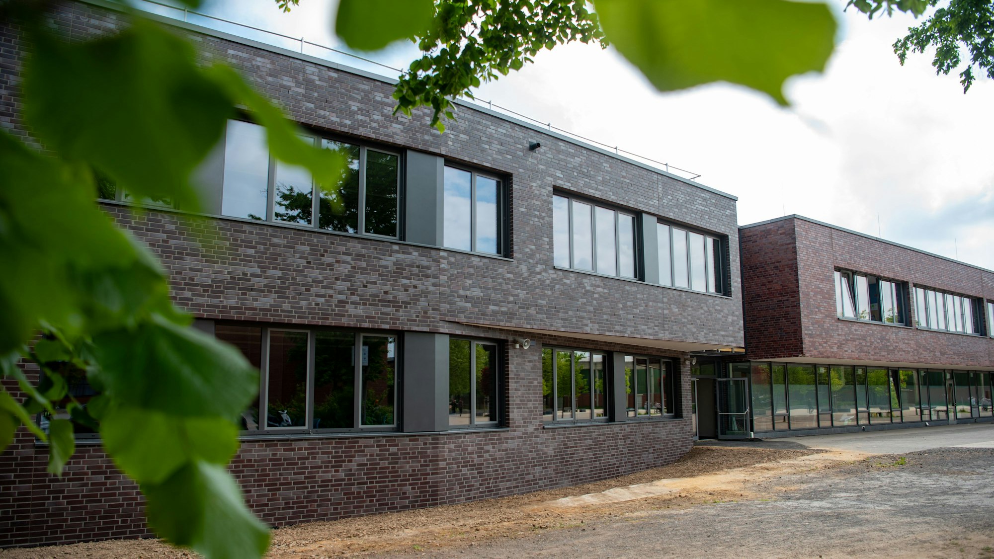 Hinter grünen Ästen sieht man ein modernes Schulgebäude mit rot-brauner Verkleidung.