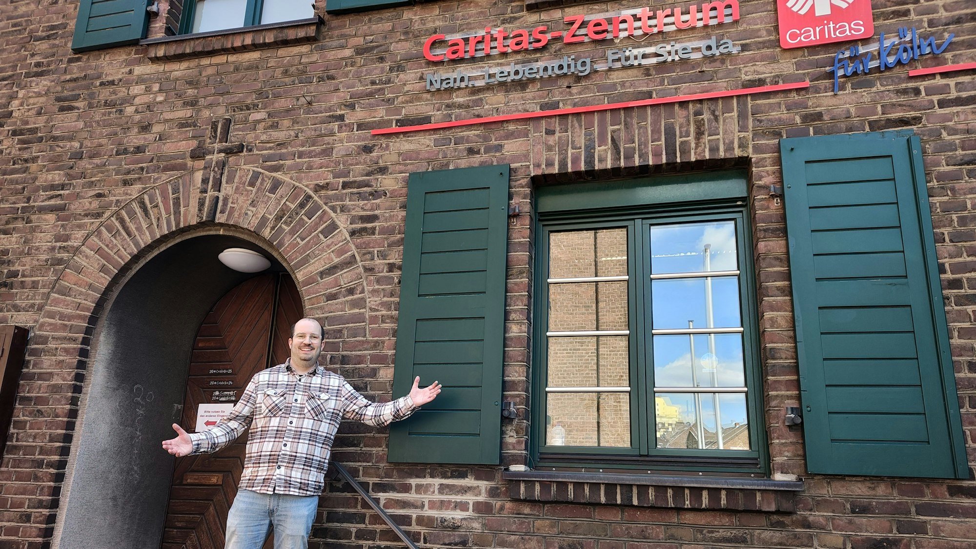 Ein Mann steht vor einem Backsteinhaus. An der Hauswand steht Caritas Zentrum Nah Lebendig Für Sie da Caritas für Köln