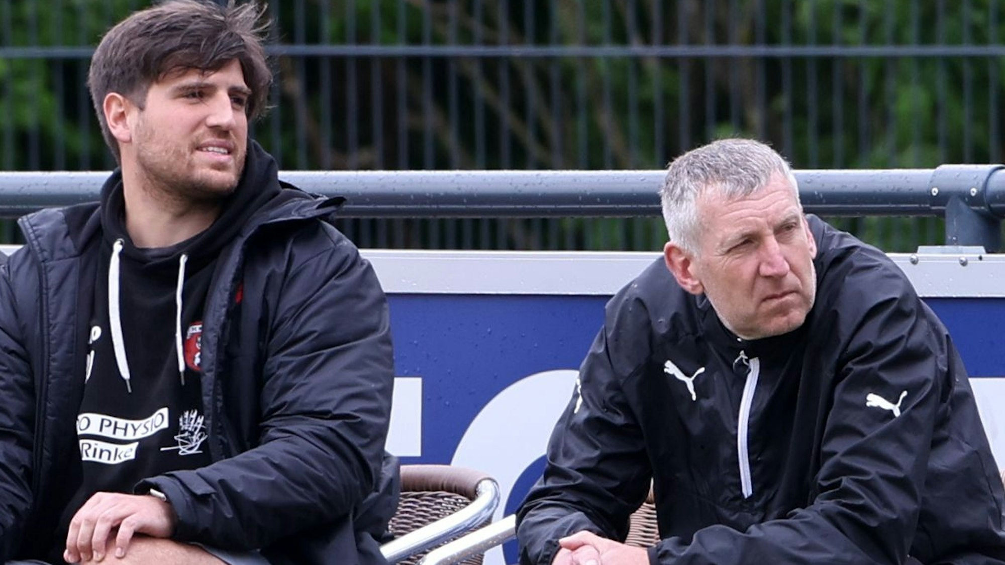 Das Foto zeigt zwei Männer, die vor einer Bande auf ihren Trainerstühlen sitzen.