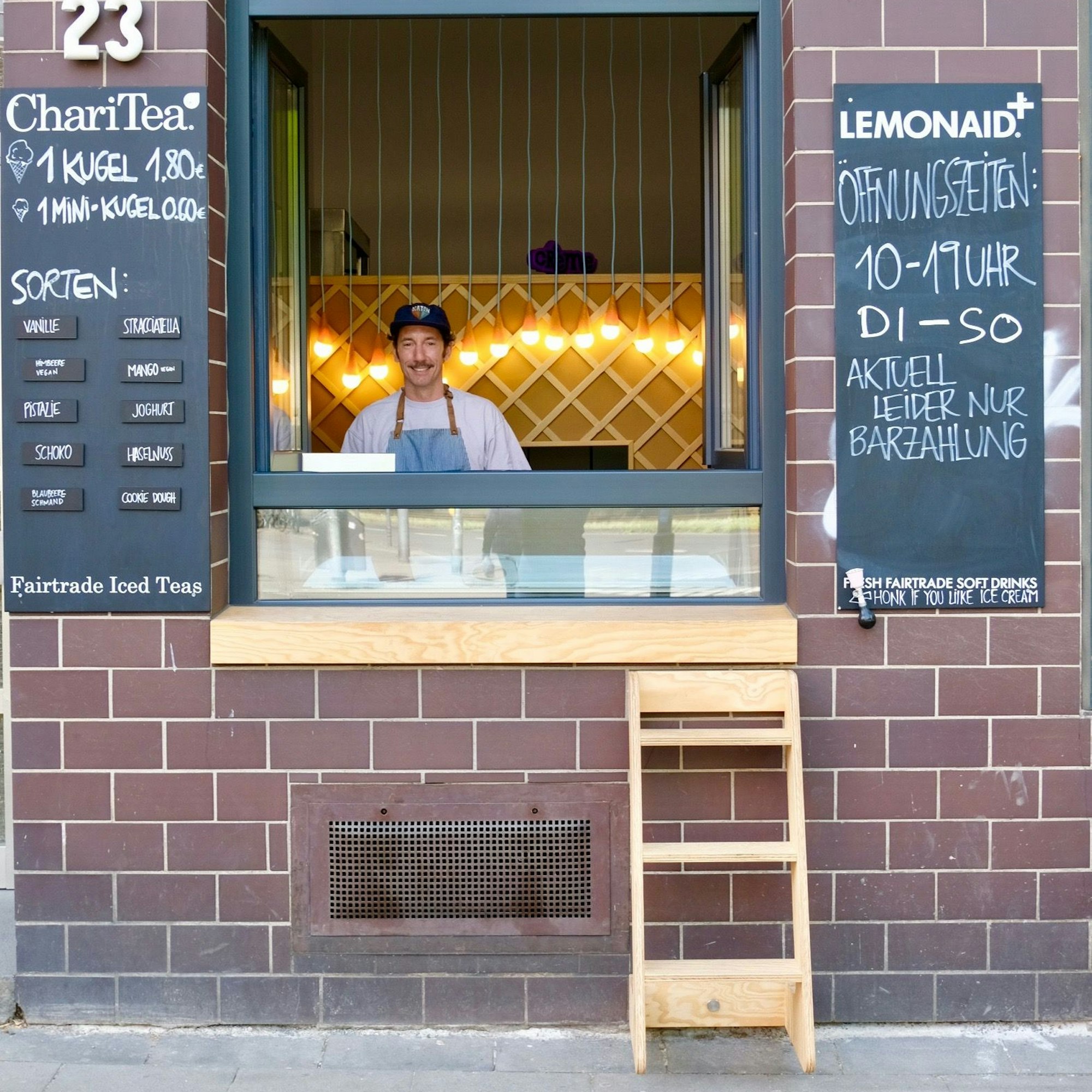 Ein Mann mit Käppi und Schnurrbart steht hinter einem offenen Verkaufsfenster. Davor steht eine kleine Holzleiter.