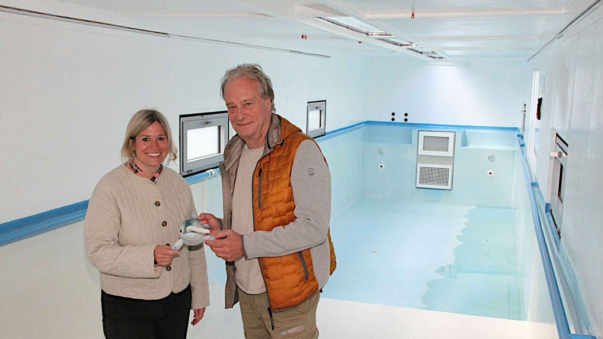 Wolfgang Schmitz vom Kreissportbund Düren und Blankenheims Bürgermeisterin Jennifer Meuren stehen im Schwimmbad-Container. Sie halten das Narwali-Maskottchen in der Hand.