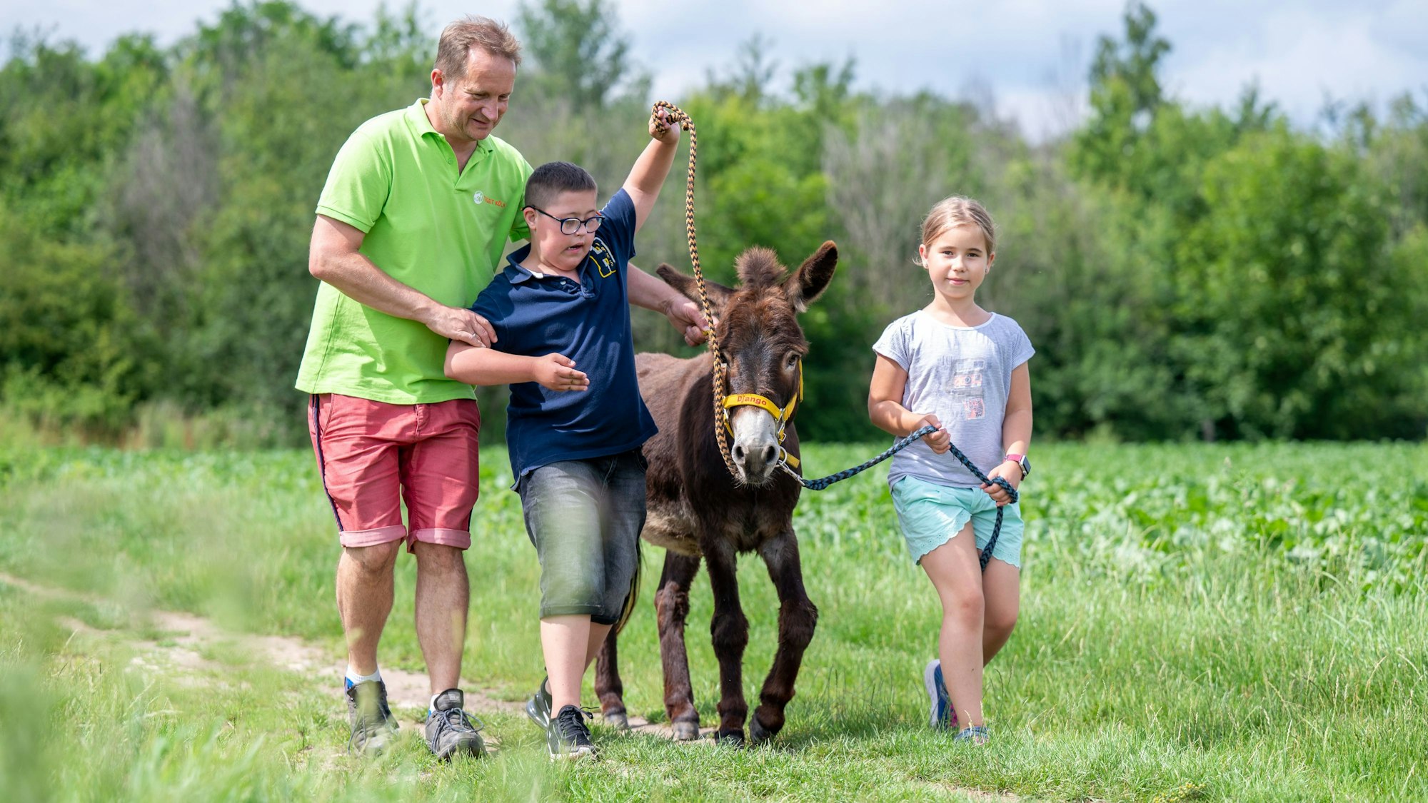 Zwei Kinder gehen mit einem Mann und einem Esel am Strick über einen Feldweg.