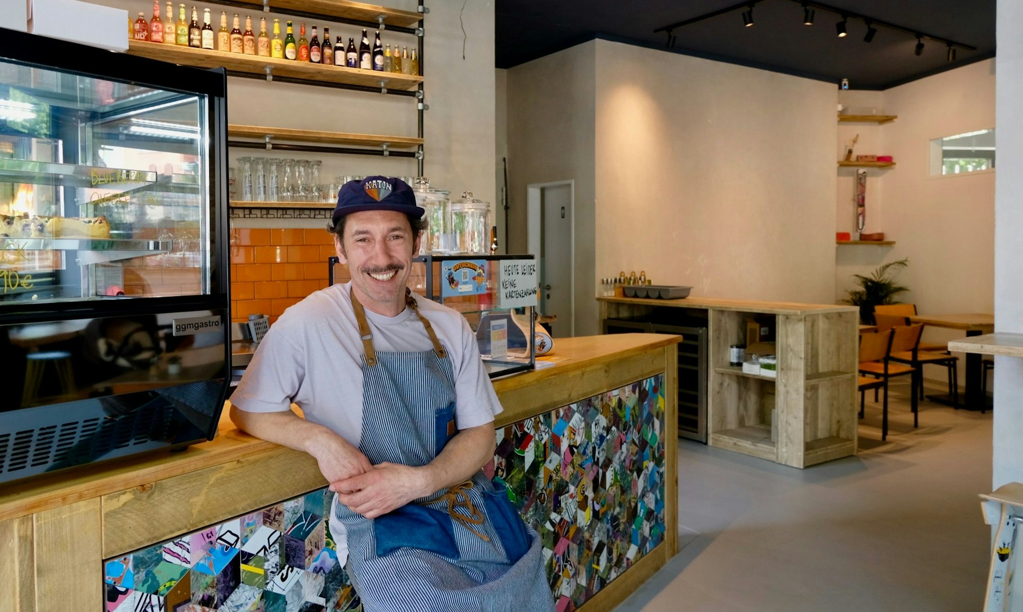 Ein Mann mit blauem Käppi und Schnauzbart steht an eine Theke gelehnt in einem Lokal.