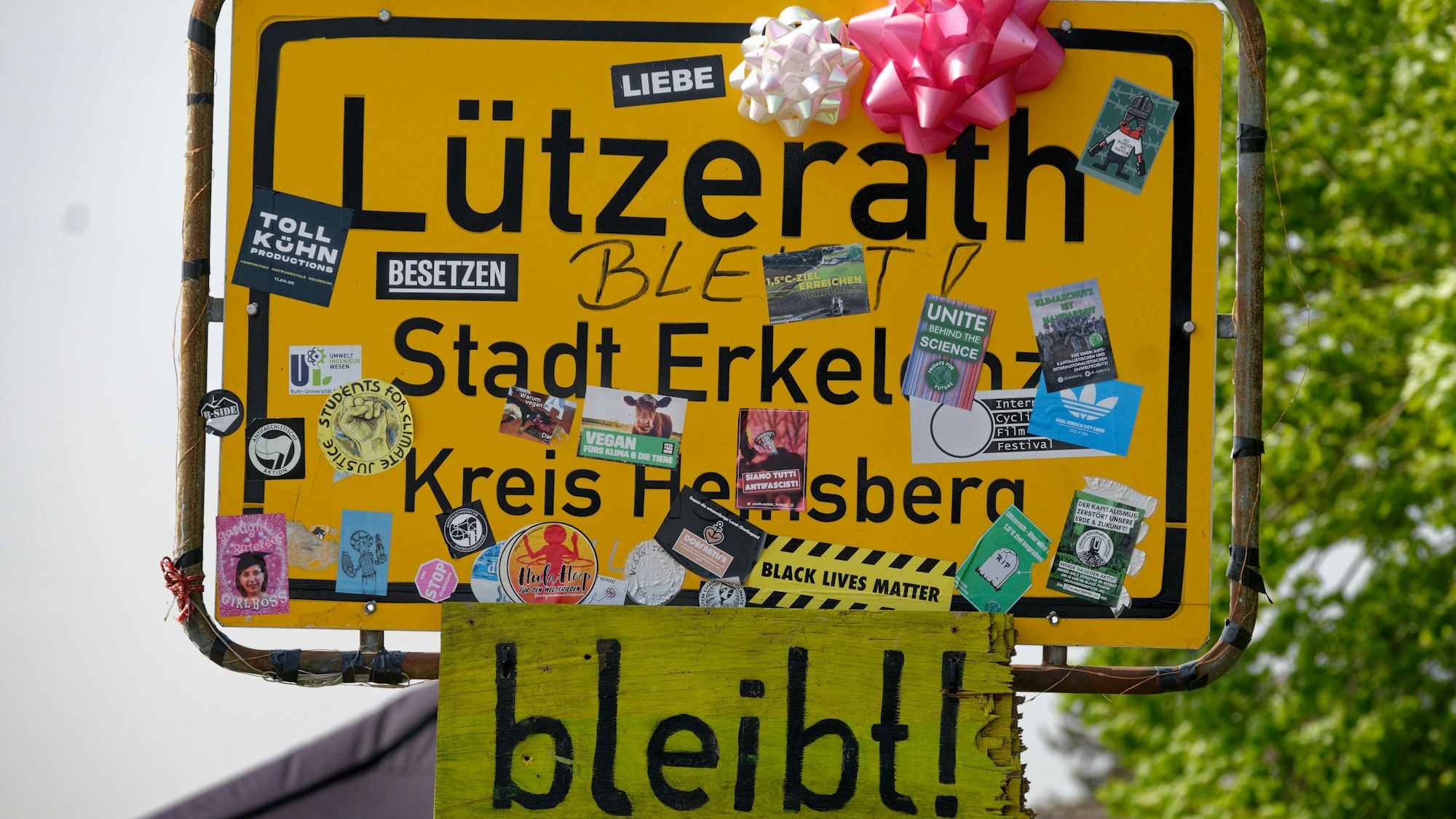 Unter dem mit Aufklebern und Schleifen versehenen Ortsschild von Lützerath hängt ein kleineres Holzschild mit der Aufschrift „bleibt!“.