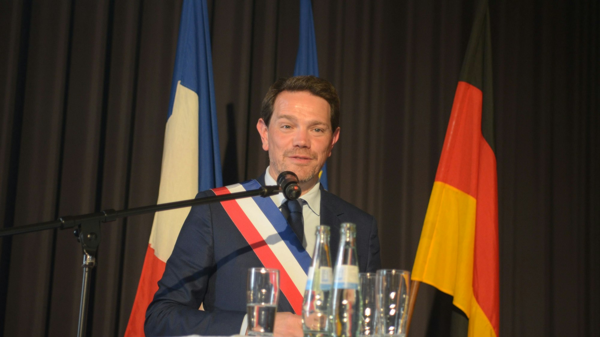 Ein Mann mit Schärpe in den französischen Nationalfarben spricht in ein Mikrofon, hinter ihm die Flaggen Deutschlands und Frankreichs.