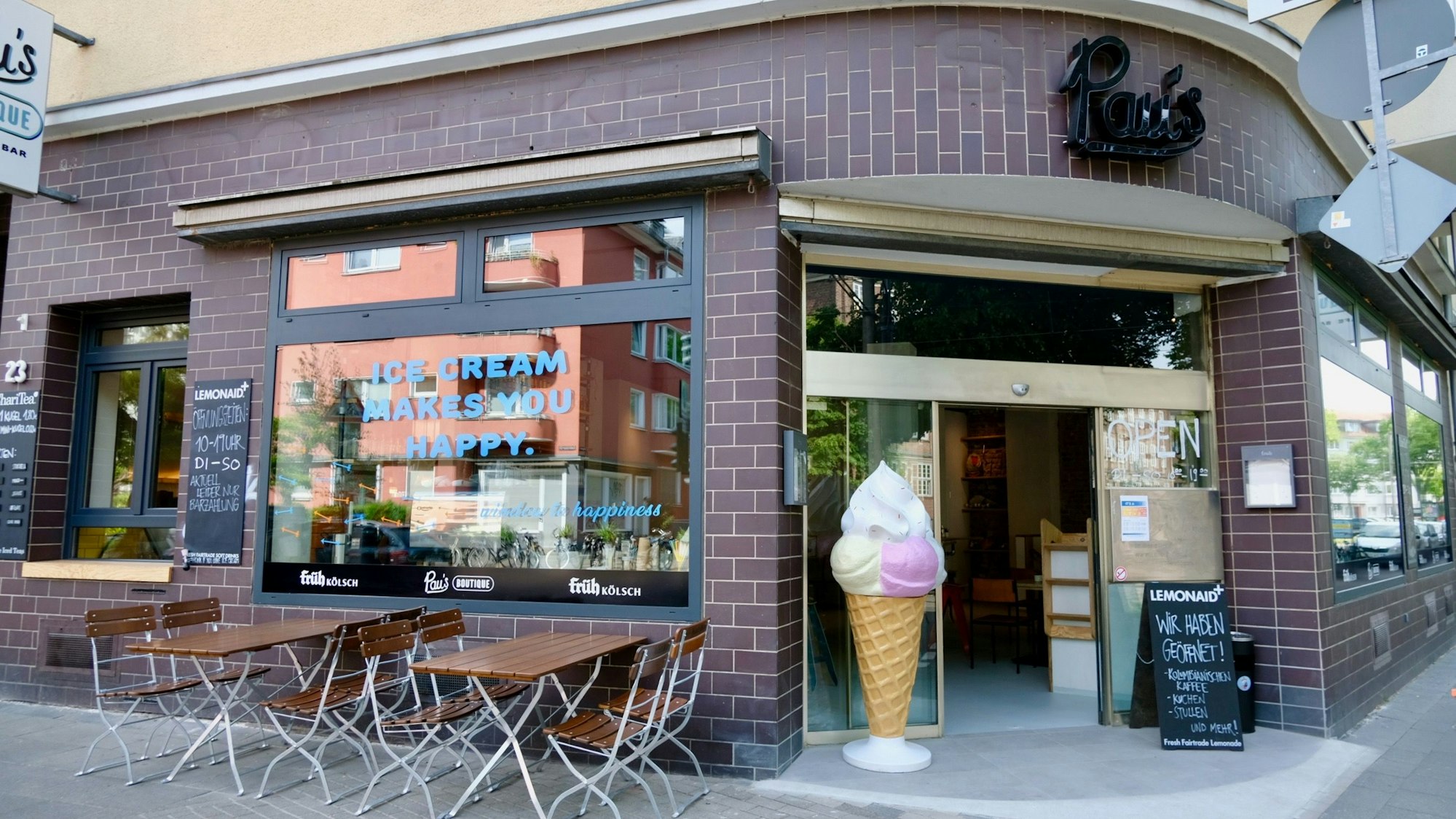 Blick auf ein Eck-Ladenlokal mit mannshoher Eiswaffel neben der Tür und Tischen vor einem Fenster.