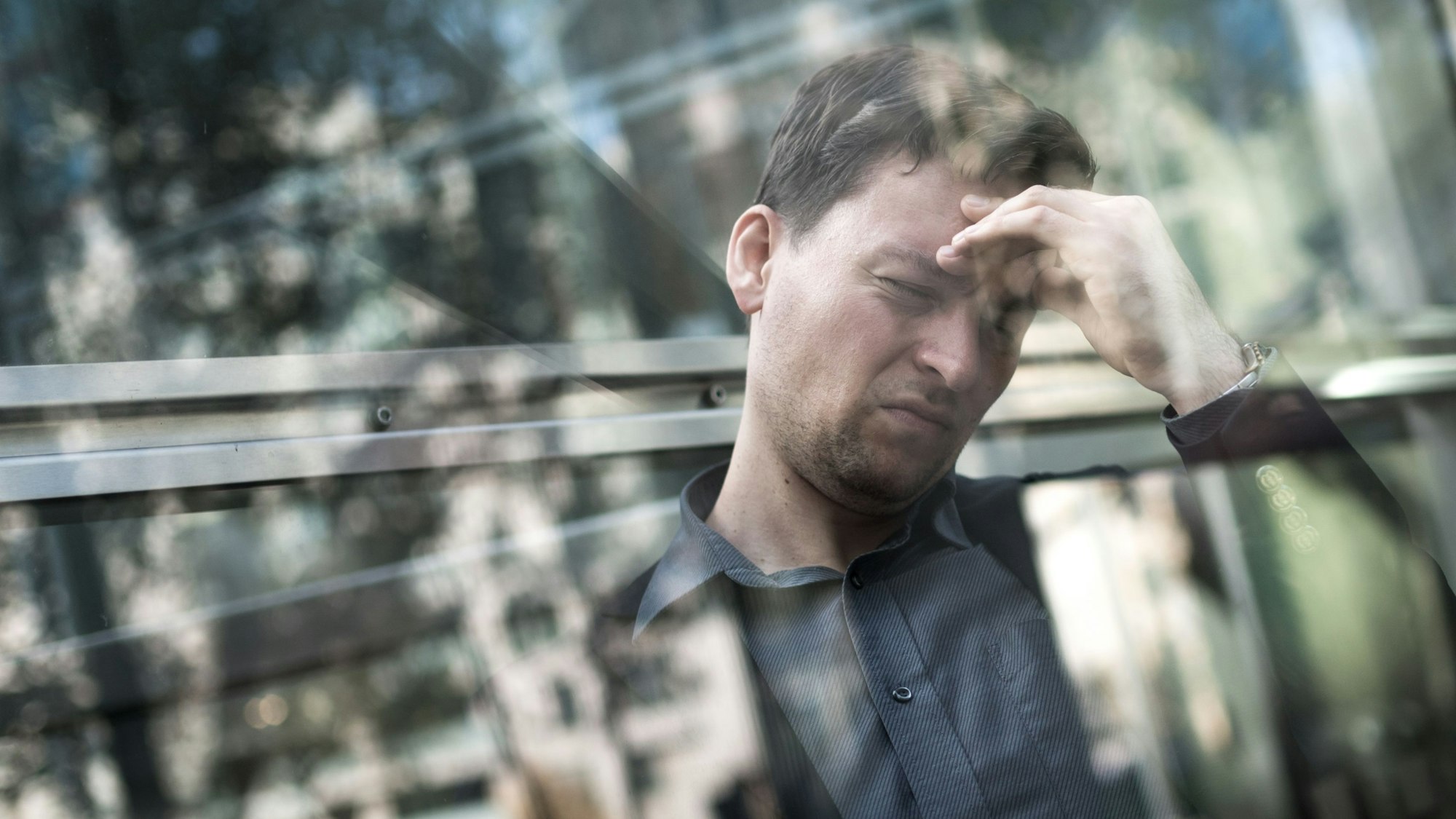 Ein Mann hält sich vor einer spiegelnden Scheibe mit schmerzverzerrtem Gesicht den Kopf.