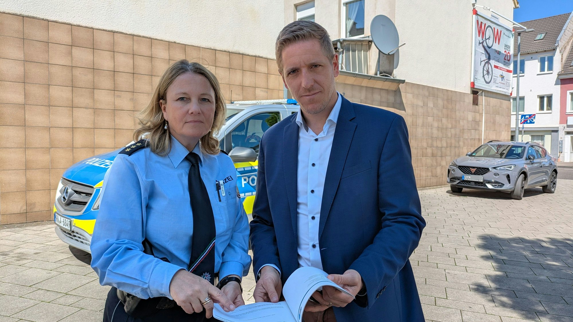 Gabriele Mälchers und Markus Ramers stehen vor einem Polizeiwagen und halten einen Stapel Papier in den Händen.